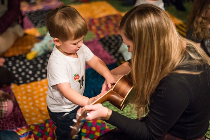 A woman playing a guitar faces a small child who is touching the guitar. They're sitting on a colourful quilt.