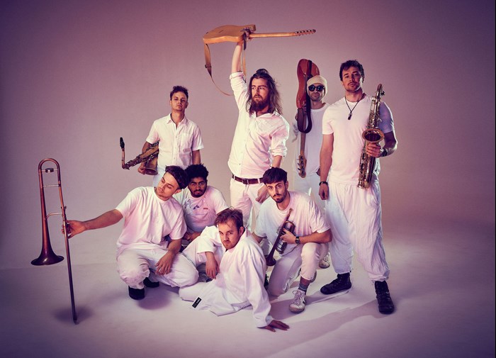 8 men pose in white outfits on a white backdrop. They are all holding different musical instruments.