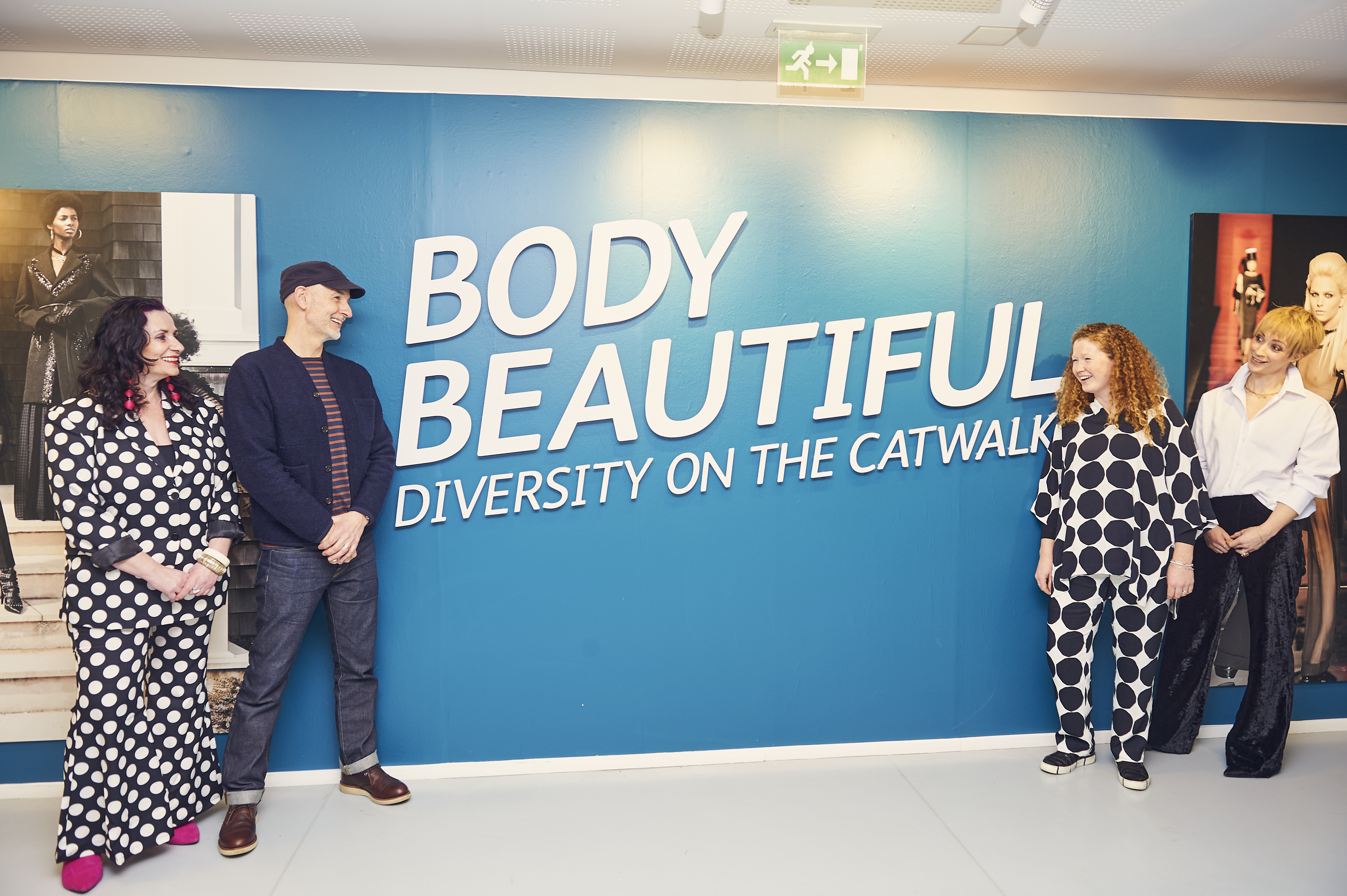 Four people pose alongside exhibition title on large wall - Body Beautiful, Diversity on the Catwalk.