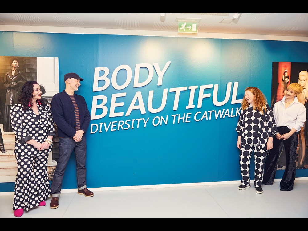 Four people pose alongside exhibition title on large wall - Body Beautiful, Diversity on the Catwalk.