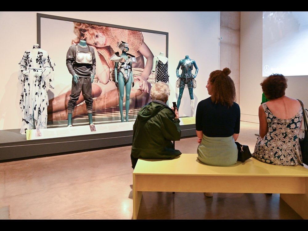 Three visitors sit on bench and look at five styled mannequins within the exhibition.