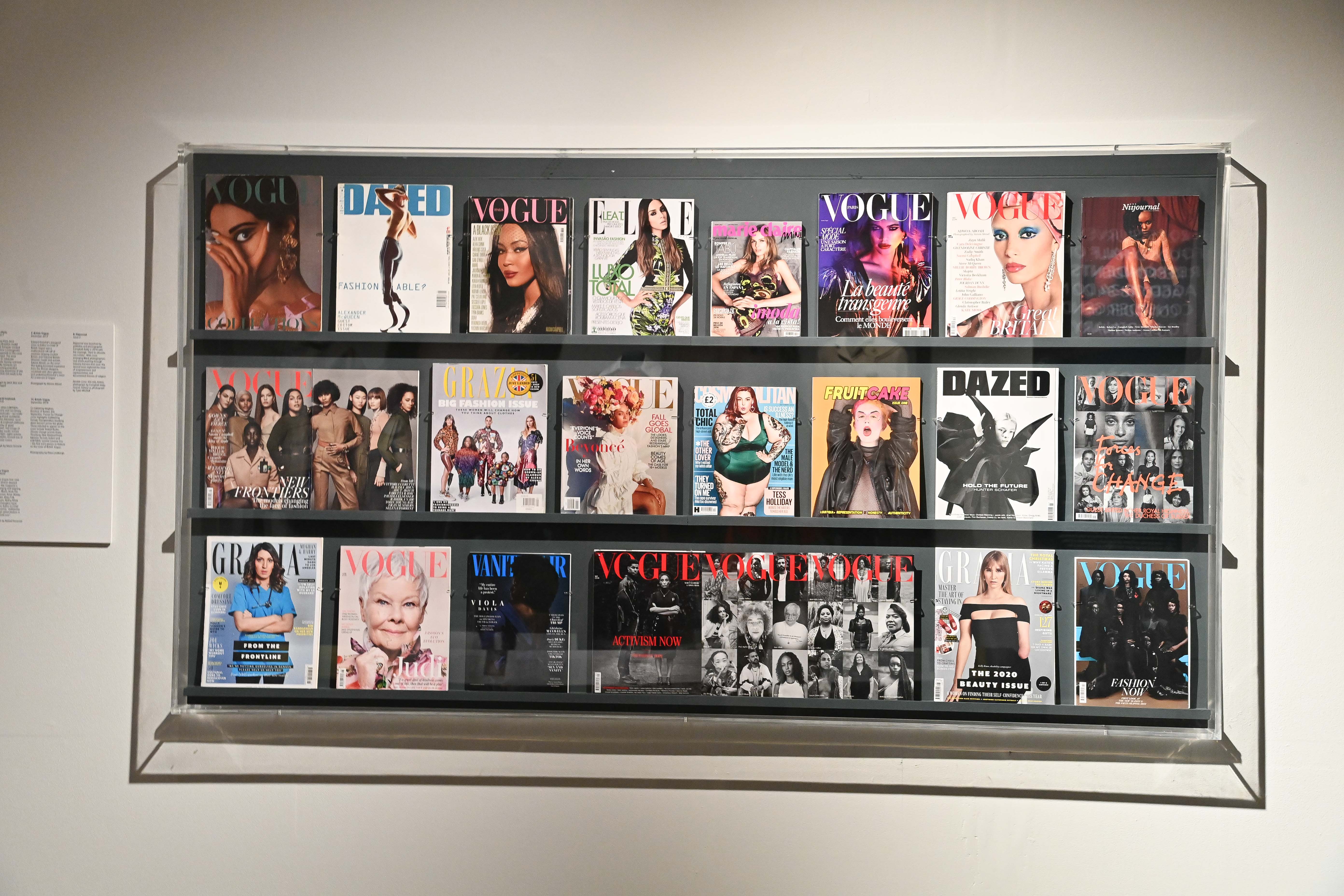 Twenty one copies of Vogue are lined on shelves within an exhibition display.