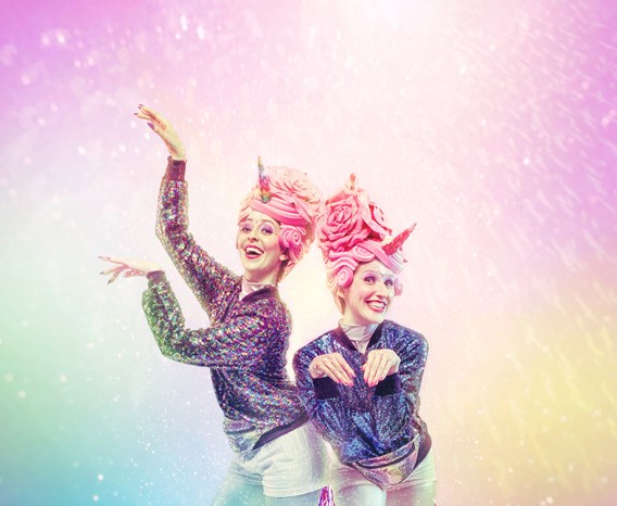 Two performers dressed and posed as unicorns with pink tails stand against a multicoloured background.