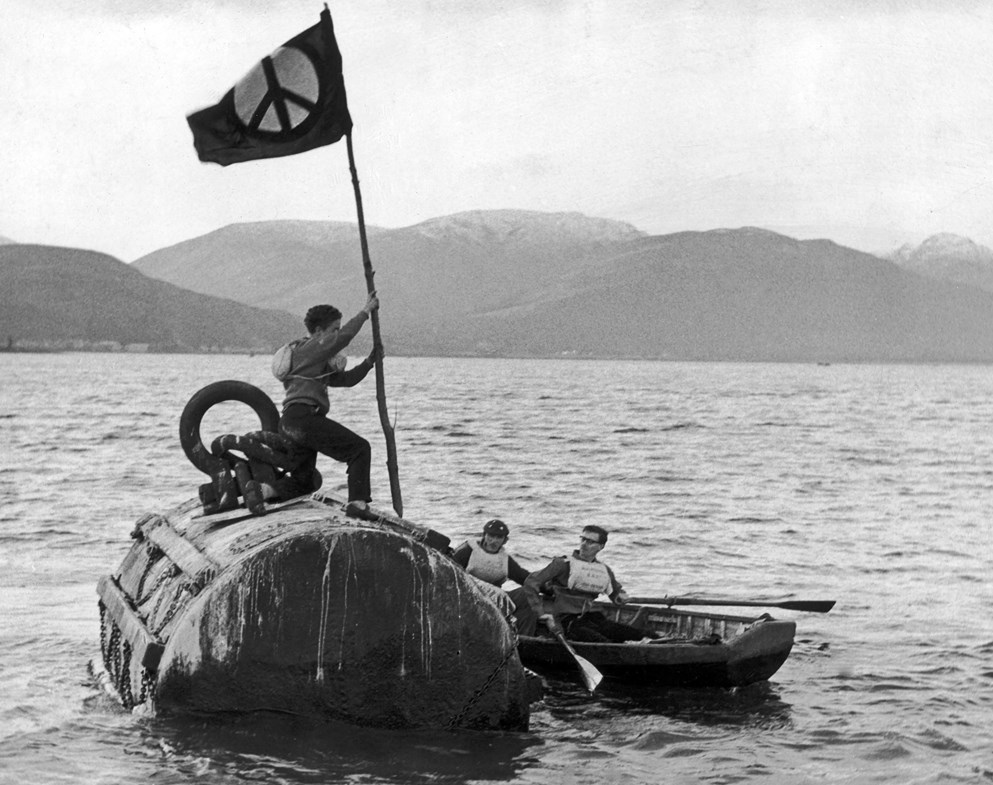 A protester stands on top of a nuclear submarine holding a peace flag in Holy Loch.