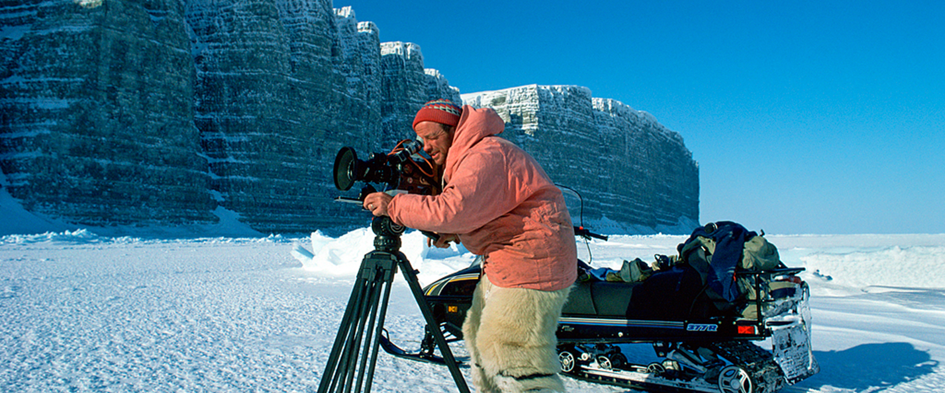 Doug Allan stands in pink jacket with camera on tripod.