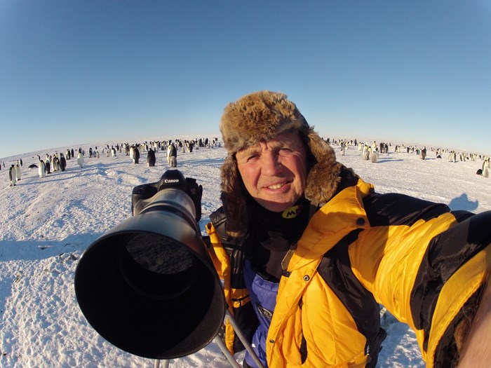 Doug Allan holds camera for a selfie with a colony of Emperor Penguins in the background.
