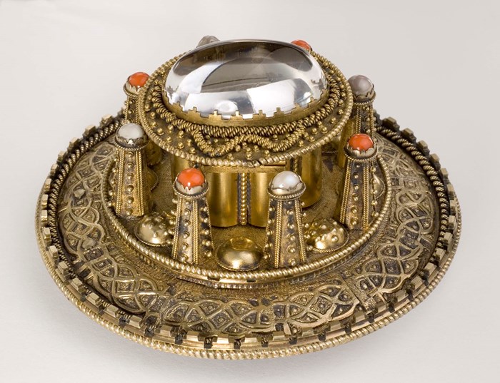 A gold coloured circular brooch with a raised centre decorated with orange and pearl jewels.