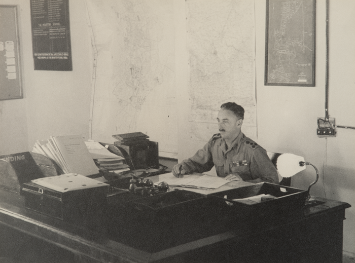 A brigadier in an office sitting at a desk with stacks of papers.