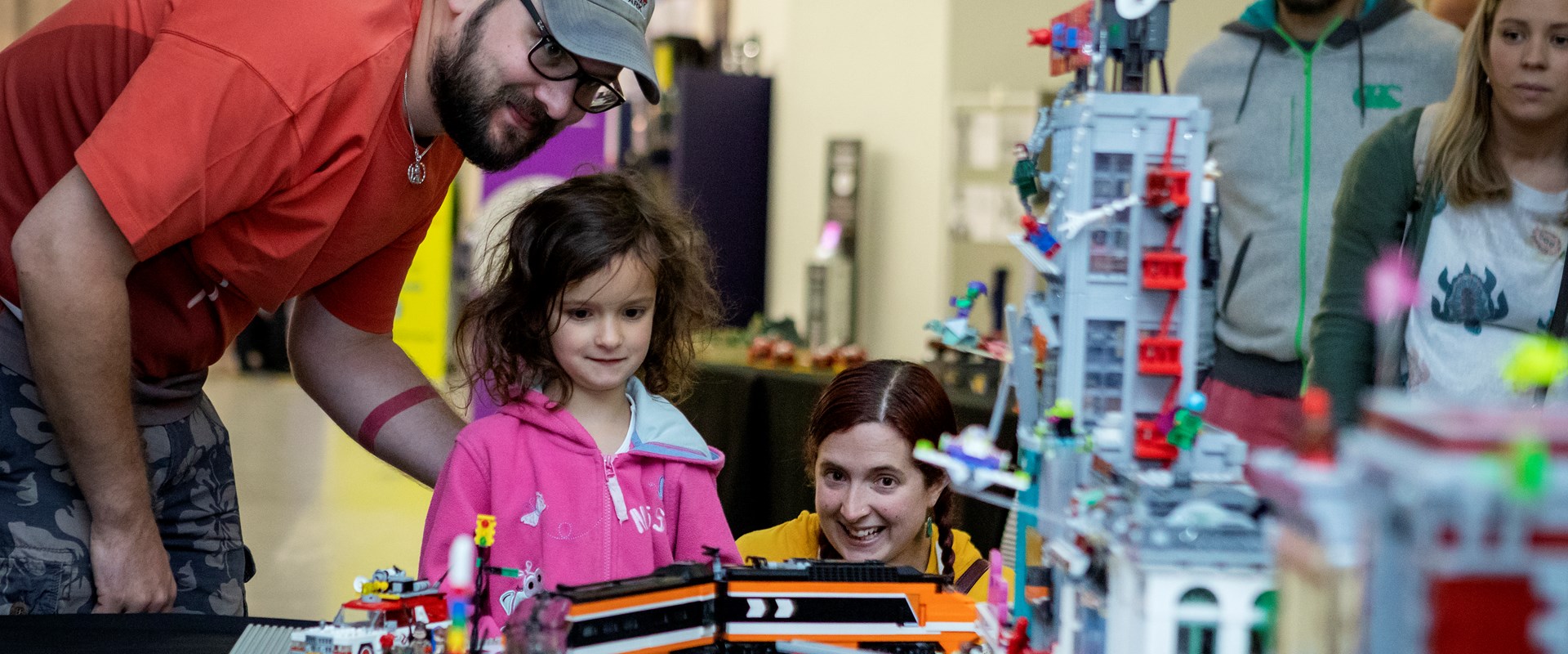 Two adults and a child look at Lego models on a table.