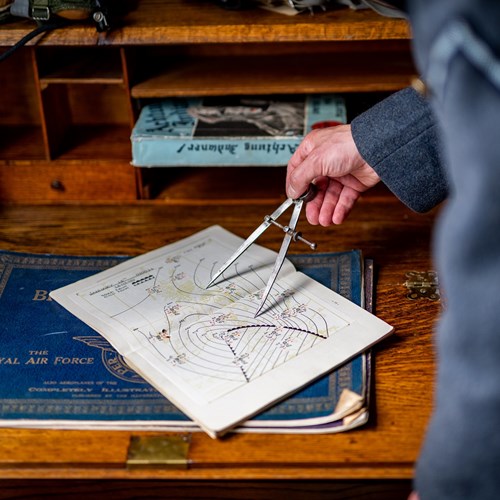 Someone is using a compass on a 1940s map. The map is placed on a desk, and the hand using the compass is visible as the person stands just out of shot, though they are wearing a blue RAF uniform.