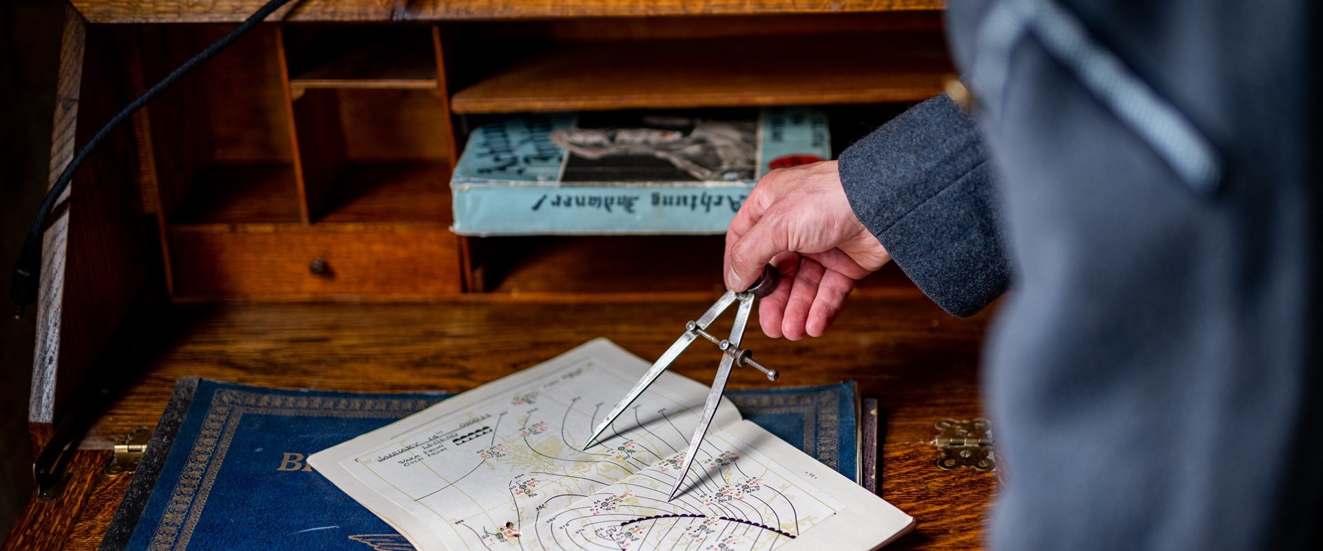 Someone is using a compass on a 1940s map. The map is placed on a desk, and the hand using the compass is visible as the person stands just out of shot, though they are wearing a blue RAF uniform.