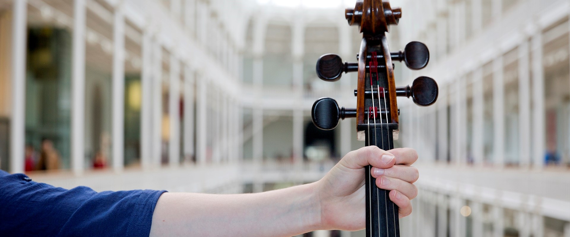 A hand holds the neck of a violin in front of the grand gallery at the National Museum of Scotland.