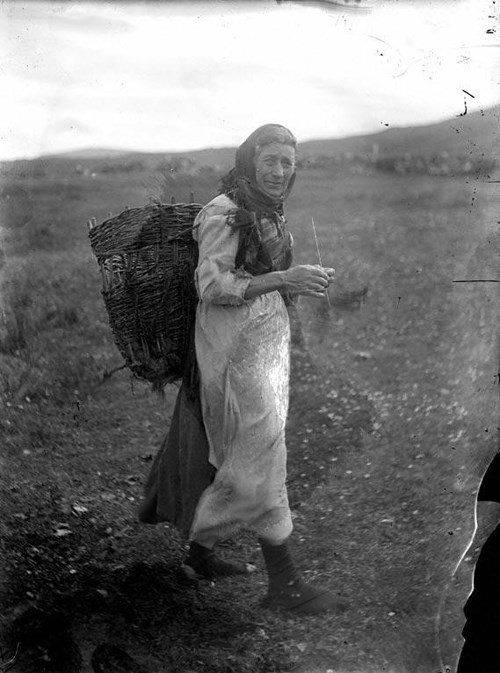 A black and white photo of a woman walking outside with a basket on her back.
