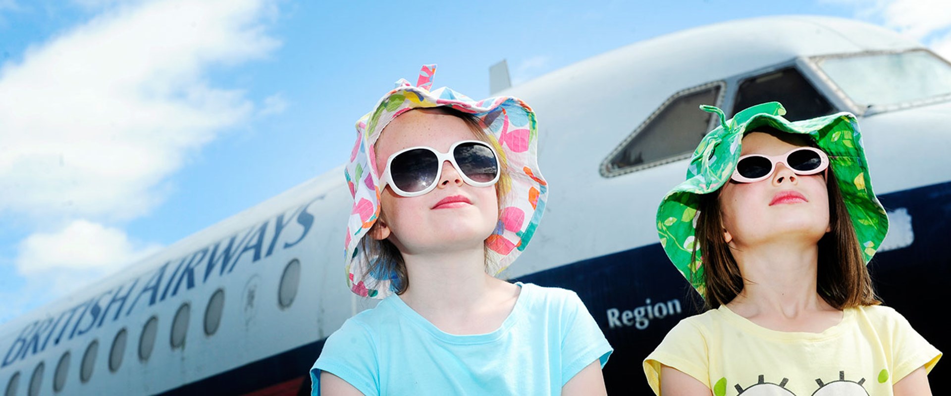Two children in sunglasses and hats look up with a British Airways plane behind them.