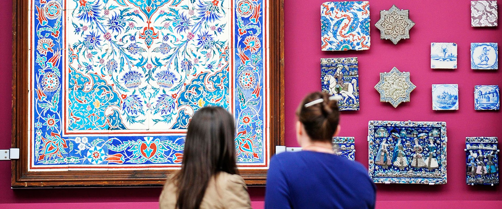 Two visitors looking at mosaic tiles hanging on a wall.