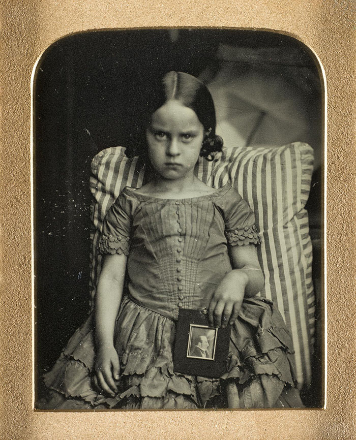 Unknown little girl sitting on a striped cushion holding a framed portrait of a man, possibly her dead father, by Ross & Thomson of Edinburgh,1847-60, ninth-plate daguerreotype.