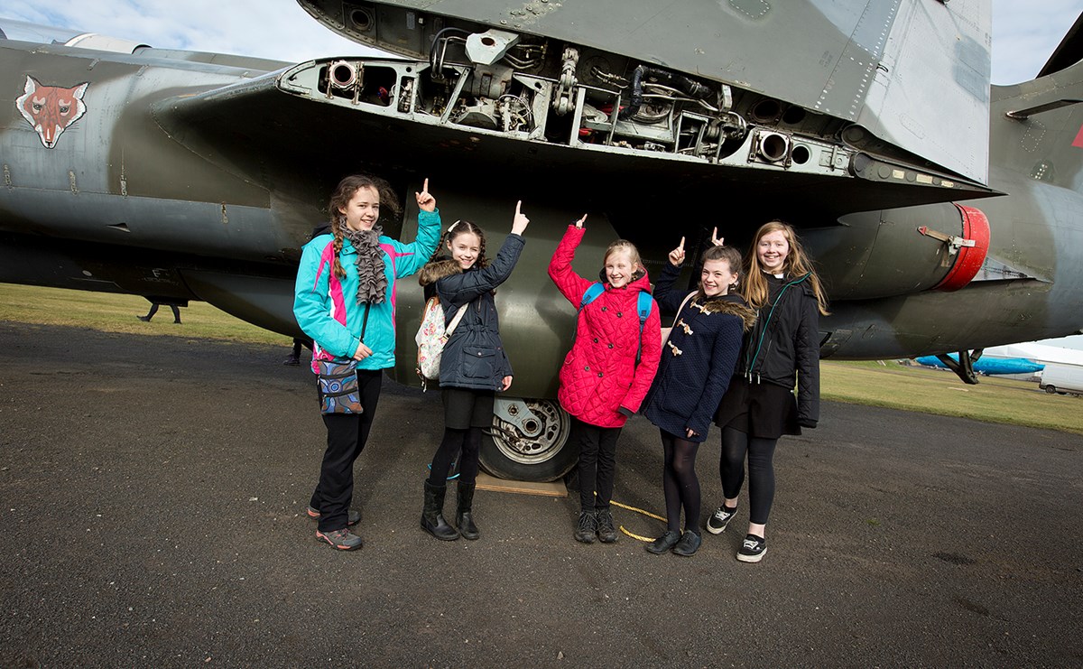 A group of children stand under the folded wing of an airplane pointing up at it