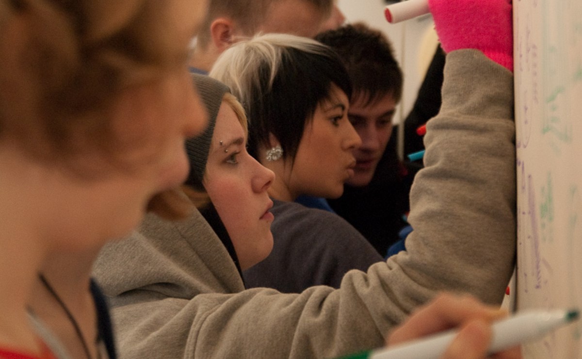 A group of 4 or 5 young people write on a whiteboard with coloured markers, looking serious and determined.