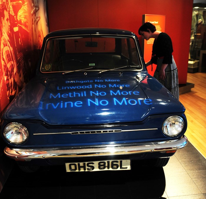 Vintage blue car in a museum gallery backed by red wallpaper and projected words on the hood reading 'Bathgate No More Linwood No More Methil No More Irvine No More'.