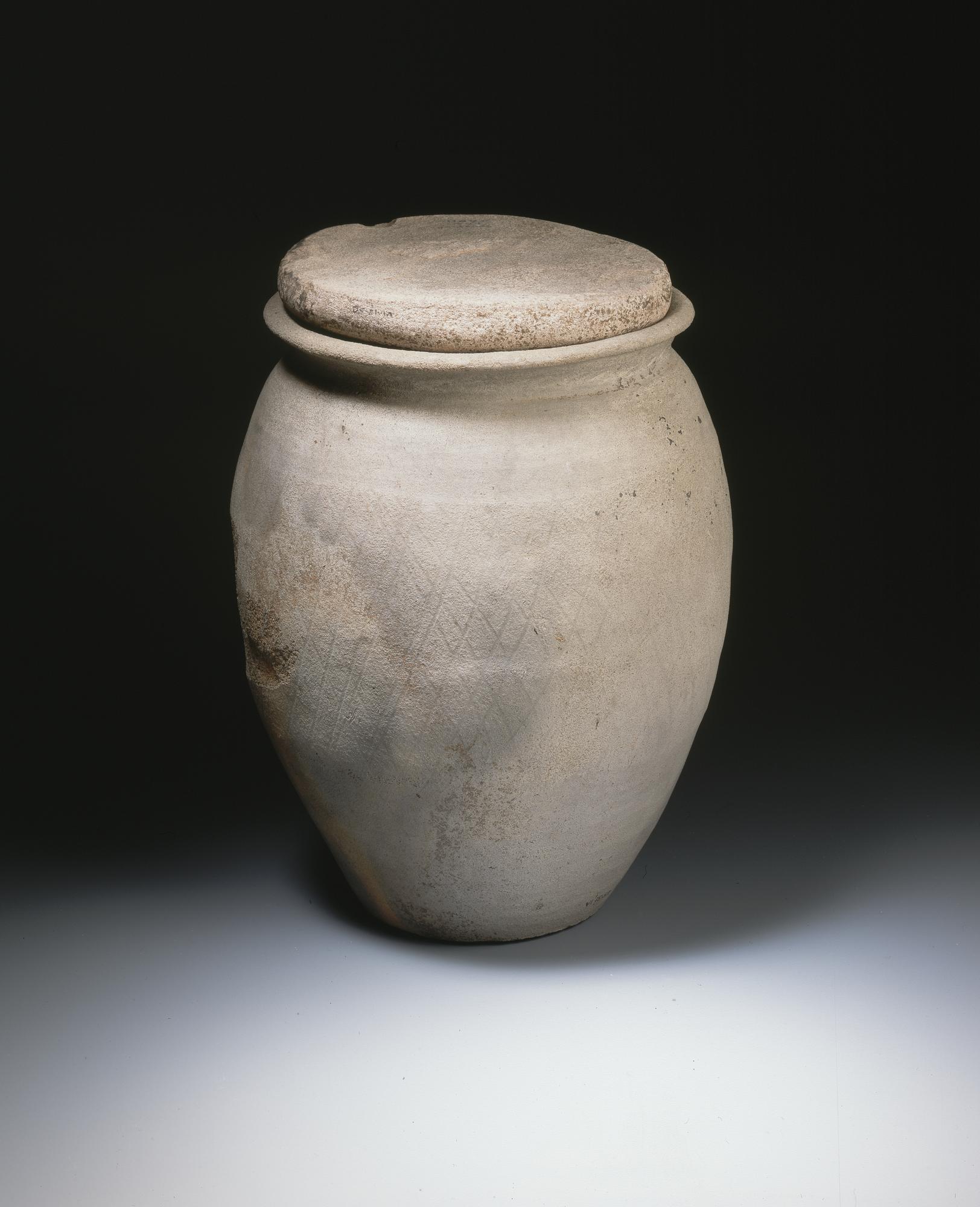 Image of Dark grey ceramic cremation urn with lattice work pattern round sides and worked stone disc as lid, from the Roman site at Newstead, 2nd century AD © National Museums Scotland
